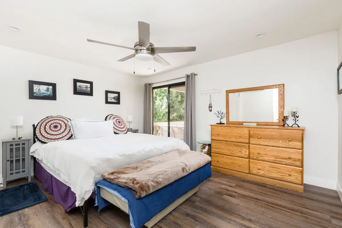 A spacious primary bedroom with a large king bed adorned with plush bedding, a wooden dresser, and framed photos on the wall. Soft natural light filters through a window with curtains, enhancing the serene and inviting atmosphere.
