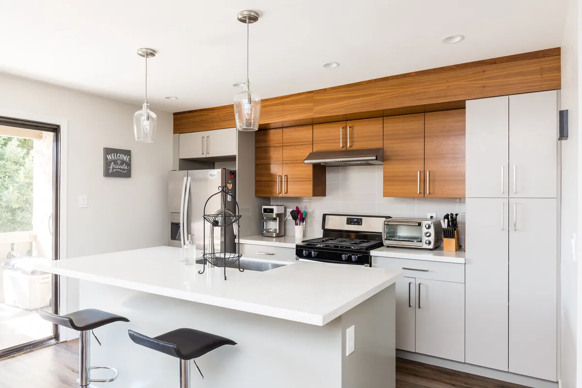 A modern kitchen with stainless steel appliances, sleek island countertop, bar stools, and functional wood cabinetry. The space is set for cooking and socializing, highlighting convenience and style.