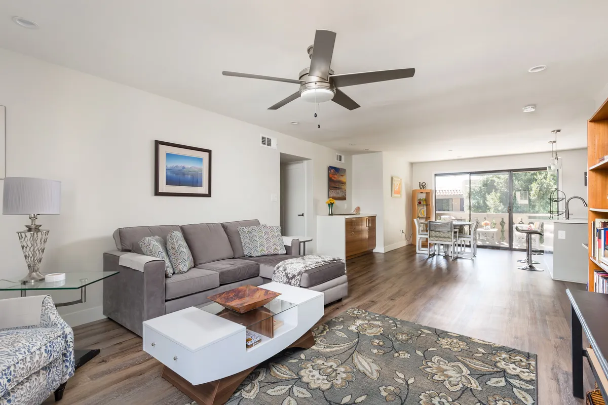 A wide-angle view of the living area shows a modern sofa, a sleek coffee table, and a dining area set against large windows that let in ample natural light. The space is open and inviting, with contemporary furniture and a minimalistic yet welcoming design, perfect for relaxing or entertaining guests.