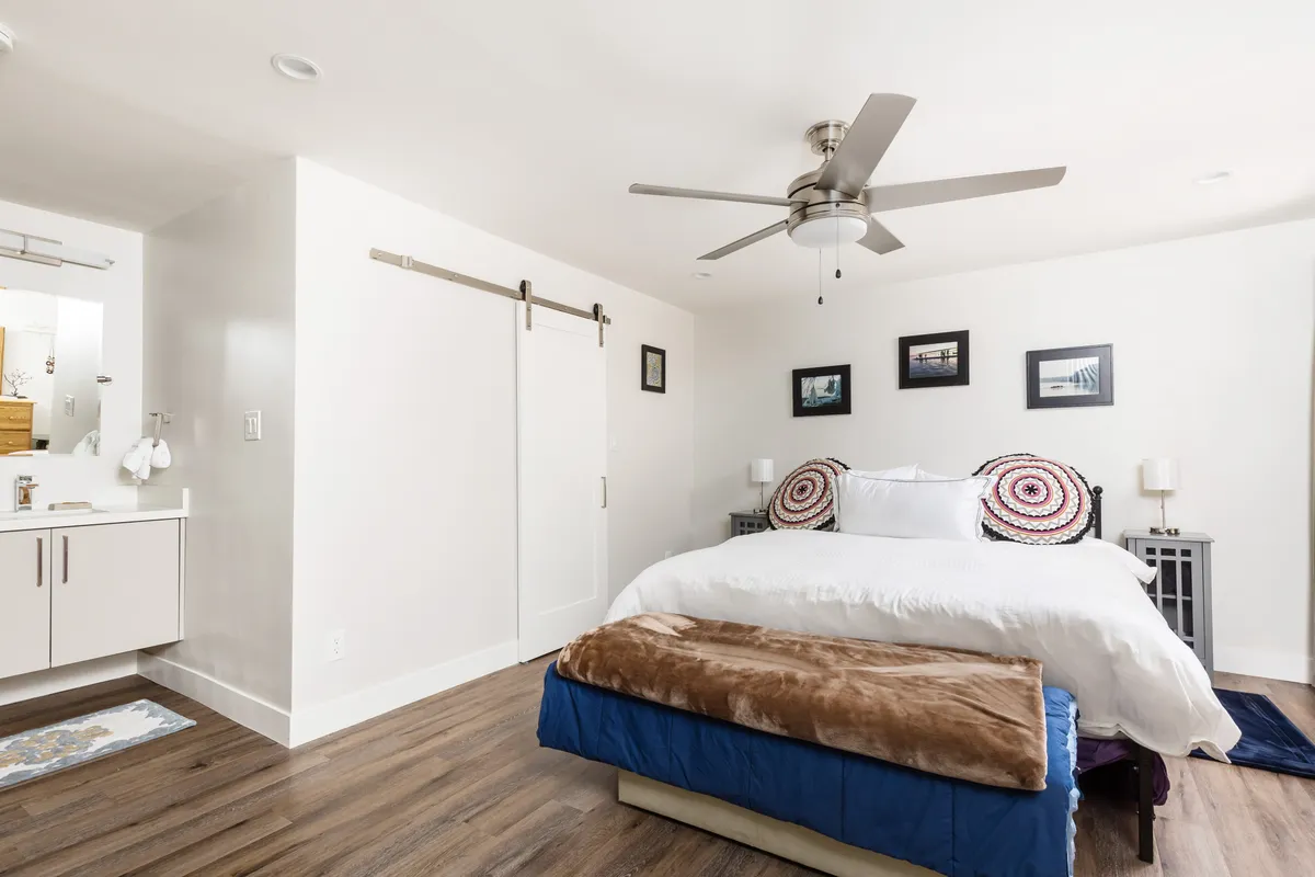 A modern king bedroom featuring a large bed with decorative pillows, a sleek ceiling fan, and framed photographs above the headboard. The room is brightened by natural light, creating a peaceful environment ideal for relaxation.