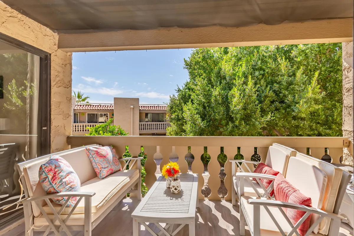The image showcases a serene outdoor patio with modern white furniture adorned with vibrant throw pillows, a stylish table with a fresh flower vase, and a backdrop of lush greenery. Morning sunlight filters softly through the trees, creating an inviting and tranquil setting perfect for relaxation or socializing.