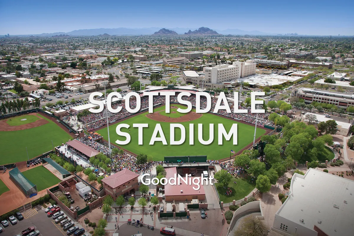 An exterior view of a vibrant baseball stadium filled with a lively crowd. The surrounding landscape includes scenic mountains, and the energy of a game day is palpable as people socialize and celebrate outdoors.