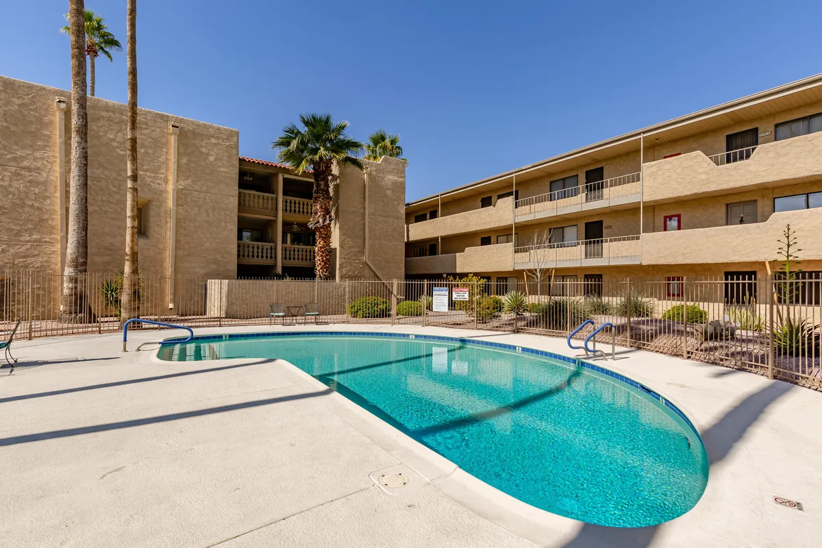A welcoming view of a community swimming pool framed by tall palm trees and the building's facade in the background. The pool area is designed for both relaxation and social interaction, with open spaces for sunbathing and gathering.